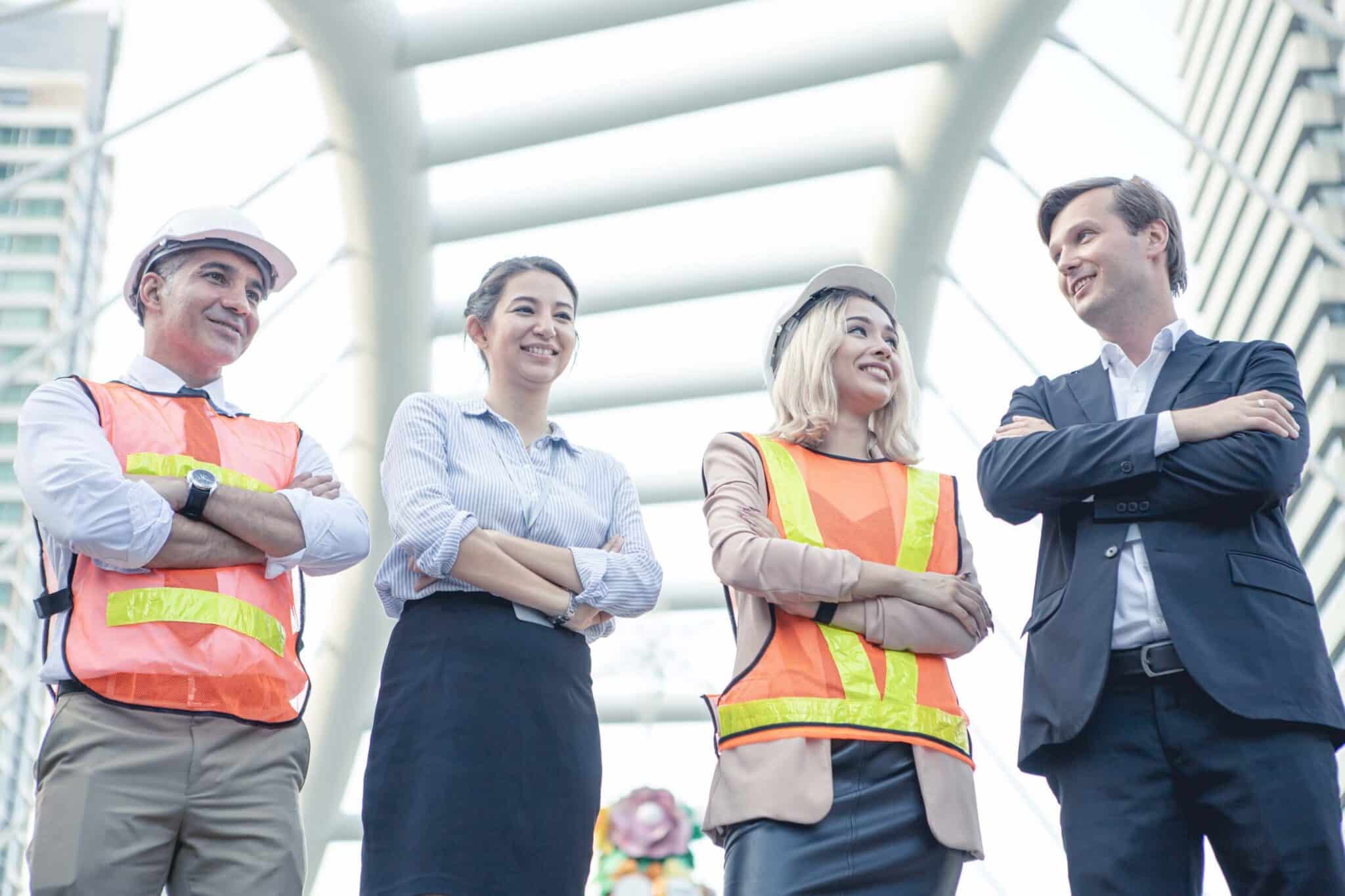 Four professionals—two in safety vests and helmets, two in business attire—stand outdoors, smiling and interacting under a modern architectural structure, exemplifying teamwork for projects requiring DCAA compliant time tracking.