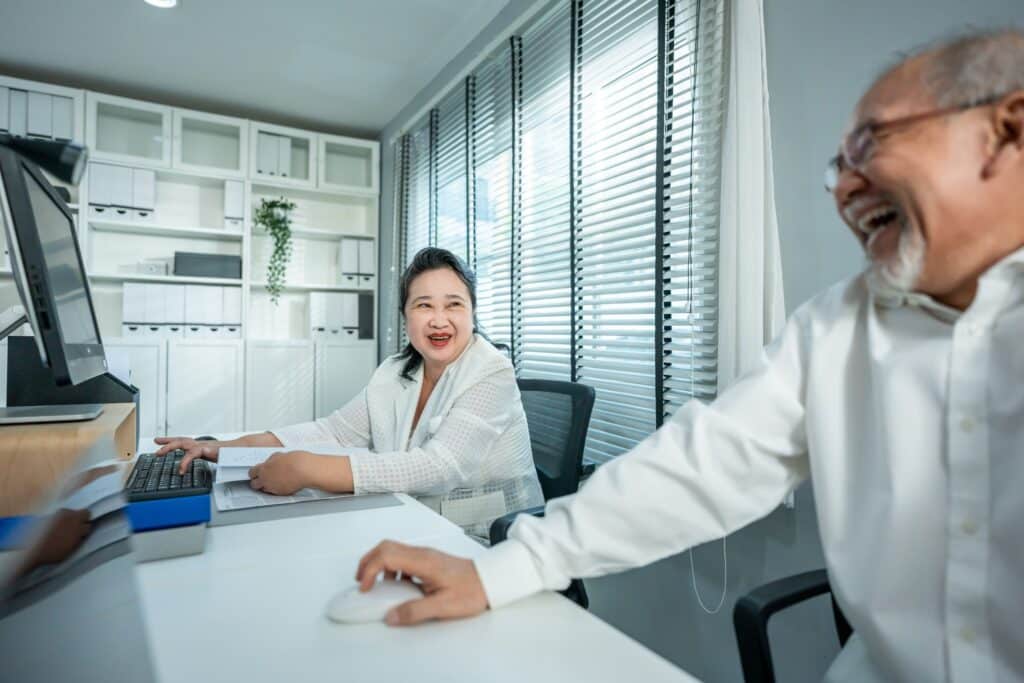 Two older adults sit at a desk in a bright office, smiling and laughing together. One is using a computer, while the other looks on, creating a cheerful and friendly atmosphere.