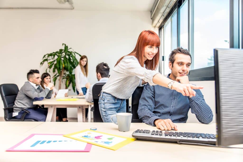 A woman stands next to a seated man, smiling and pointing at his computer screen in a modern office. Other colleagues work and talk together at nearby desks in the bright, open workspace.