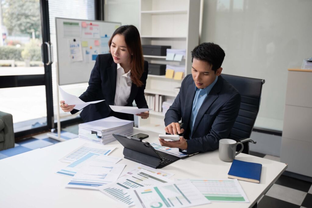 Two business professionals review documents and charts, discussing bill rate vs pay rate at a desk covered with papers, a laptop, and a coffee mug in a modern office with large windows and a whiteboard in the background.