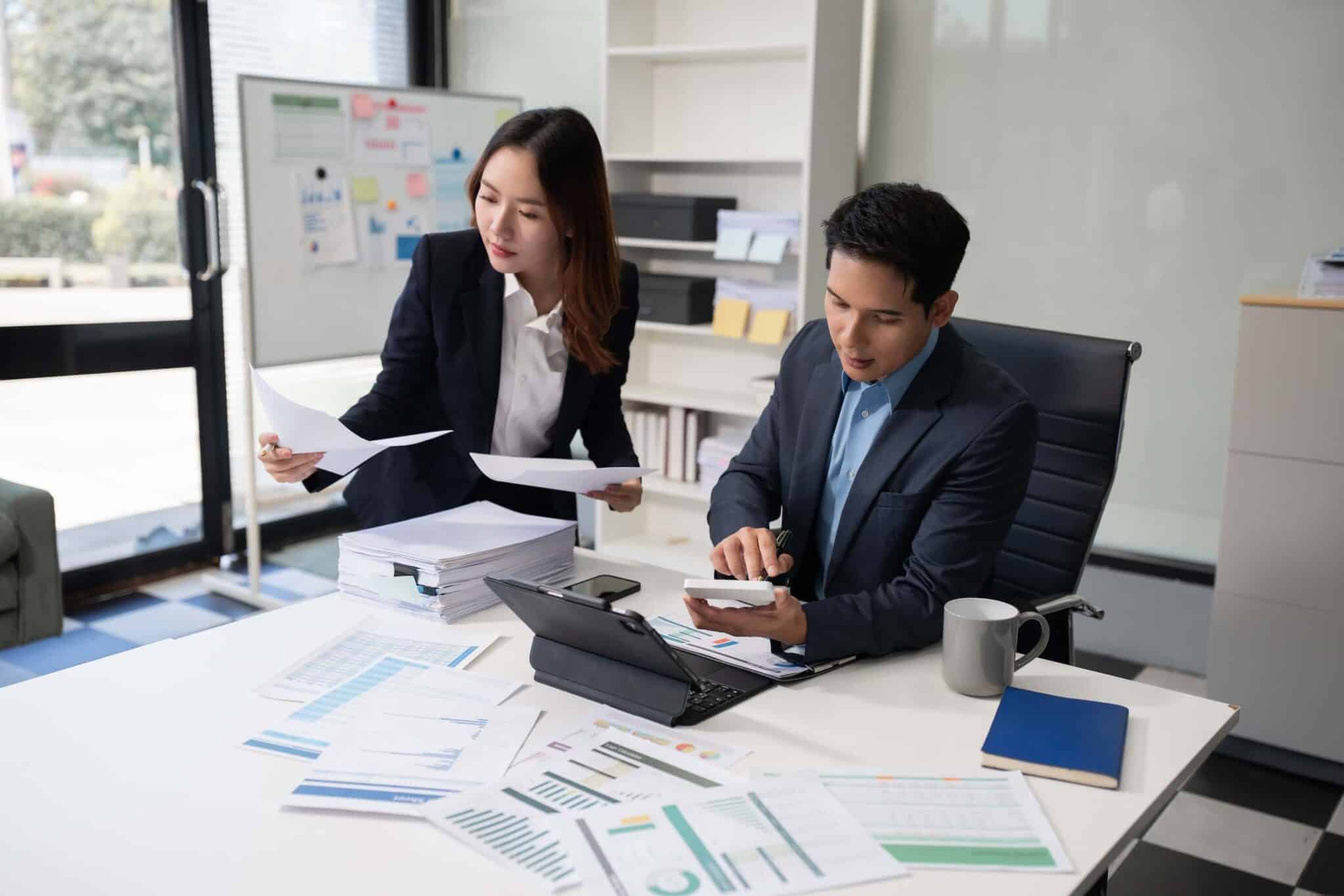 Two business professionals review documents and charts, discussing bill rate vs pay rate at a desk covered with papers, a laptop, and a coffee mug in a modern office with large windows and a whiteboard in the background.