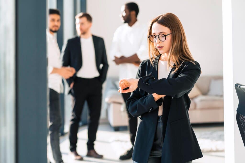 A woman in business attire checks her watch, reflecting on time tracking, while three men in suits and business casual talk in the background of a brightly lit office space.