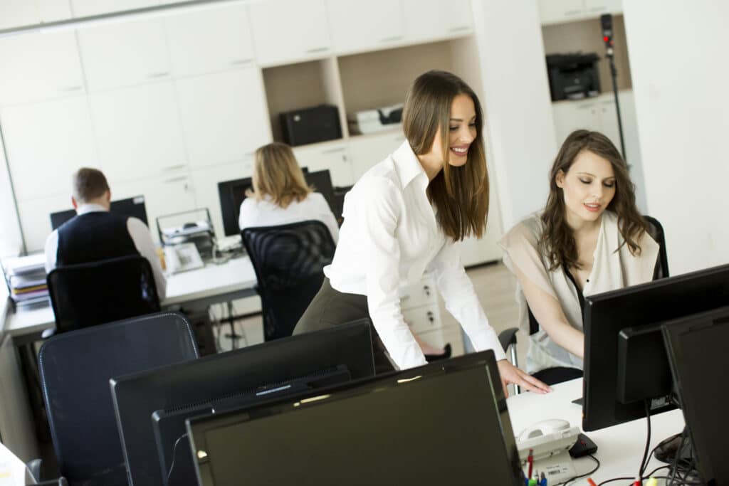 Two women smiling and working together at a computer in a bright office, with two other people working at desks in the background. Computer monitors and office equipment are visible.