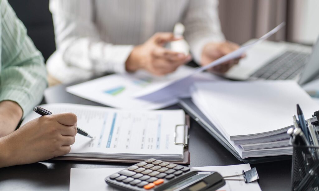 Two people work at a desk with financial documents, a laptop, and a calculator, discussing bill rate vs pay rate. One writes on a clipboard while the other holds papers, suggesting a meeting about finances and compensation.