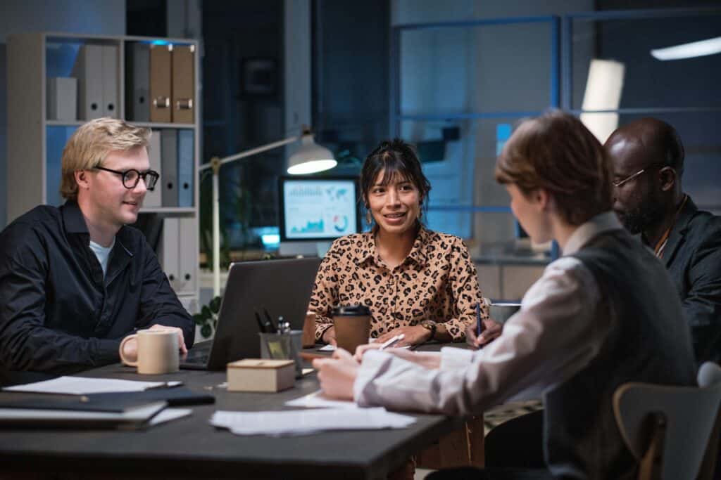 Four people sit around a table in an office, discussing time tracking solutions. Papers, a laptop, and coffee cups are on the table. Shelves and a lamp in the background create a collaborative setting for reviewing attendance software options.