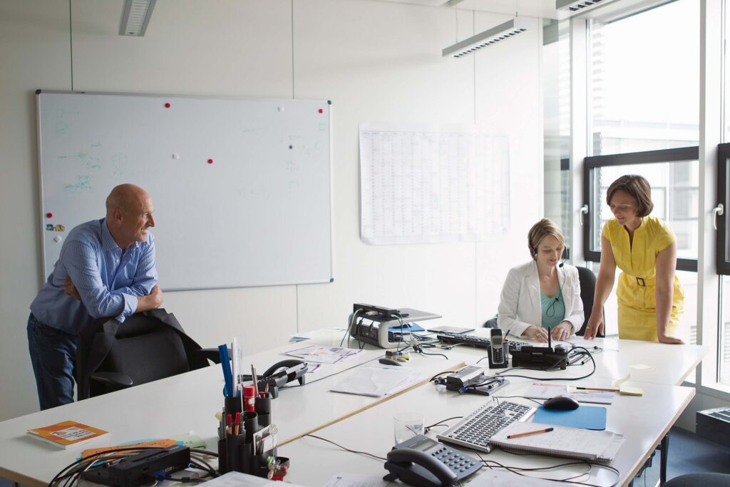 Three people are in a bright office. A man stands by a whiteboard while two women, one seated and one standing, review documents detailing the bill rate vs pay rate at a desk cluttered with papers, phones, and office supplies.