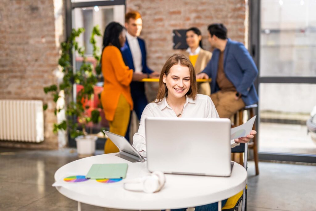 A woman smiles while working at a laptop and holding papers at a round table in a modern office, possibly reviewing the best Replicon alternatives. Four colleagues chat in the background near large windows.