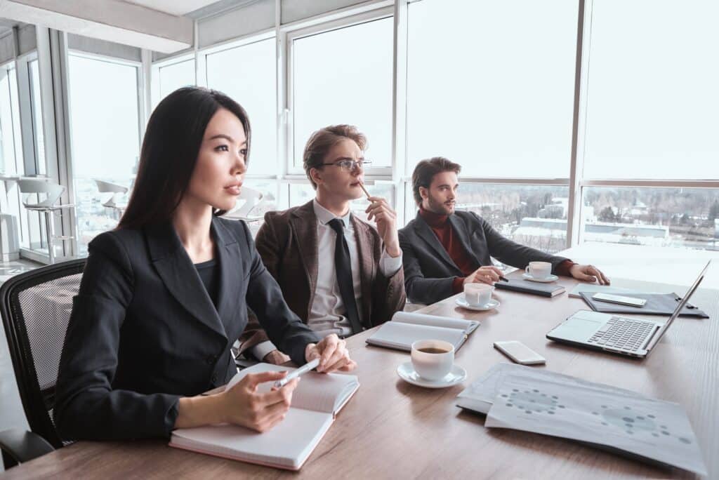 Three people in business attire sit at a conference table with notebooks, coffee cups, and a laptop, attentively listening during a meeting in a bright office with large windows.