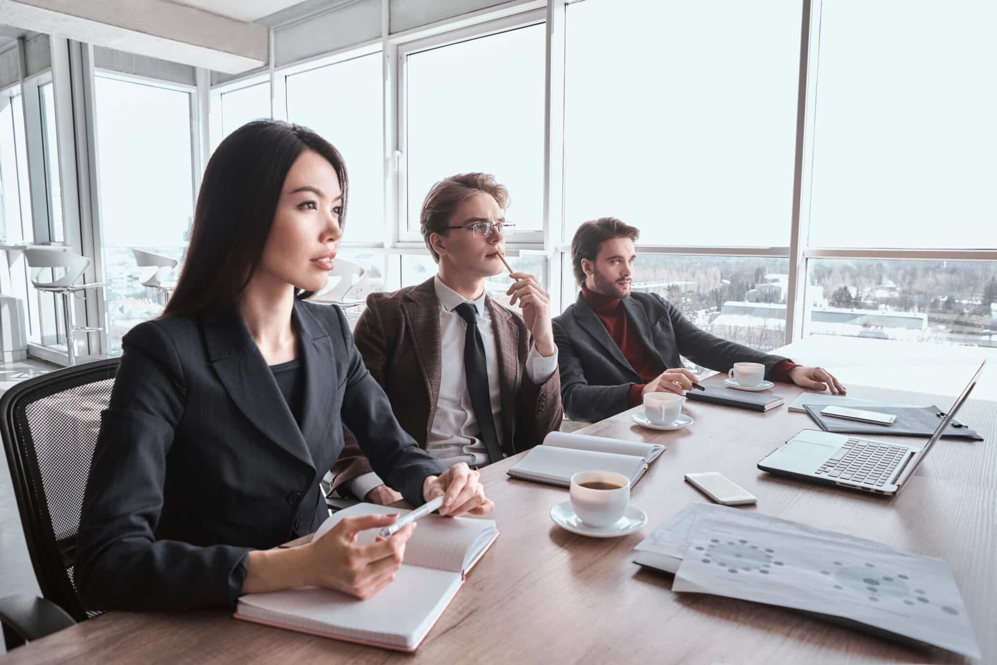Three people in business attire sit at a conference table with notebooks, coffee cups, and a laptop, attentively listening during a meeting in a bright office with large windows.