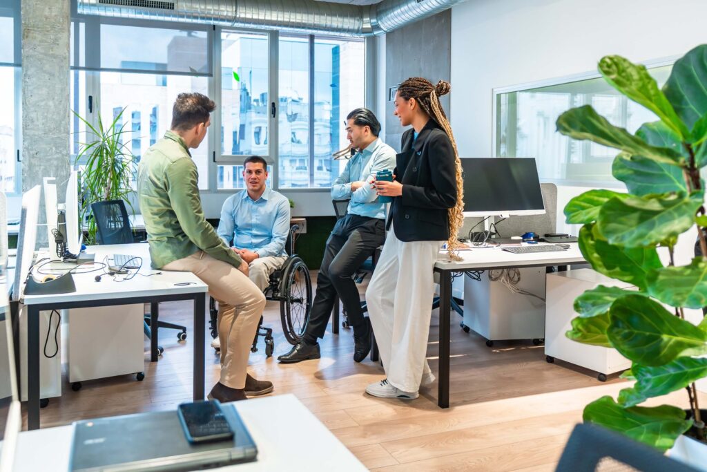 Four colleagues, including one man in a wheelchair, have a conversation in a modern office with large windows and plants. The group appears engaged and relaxed while discussing the best replicon alternatives for their team.