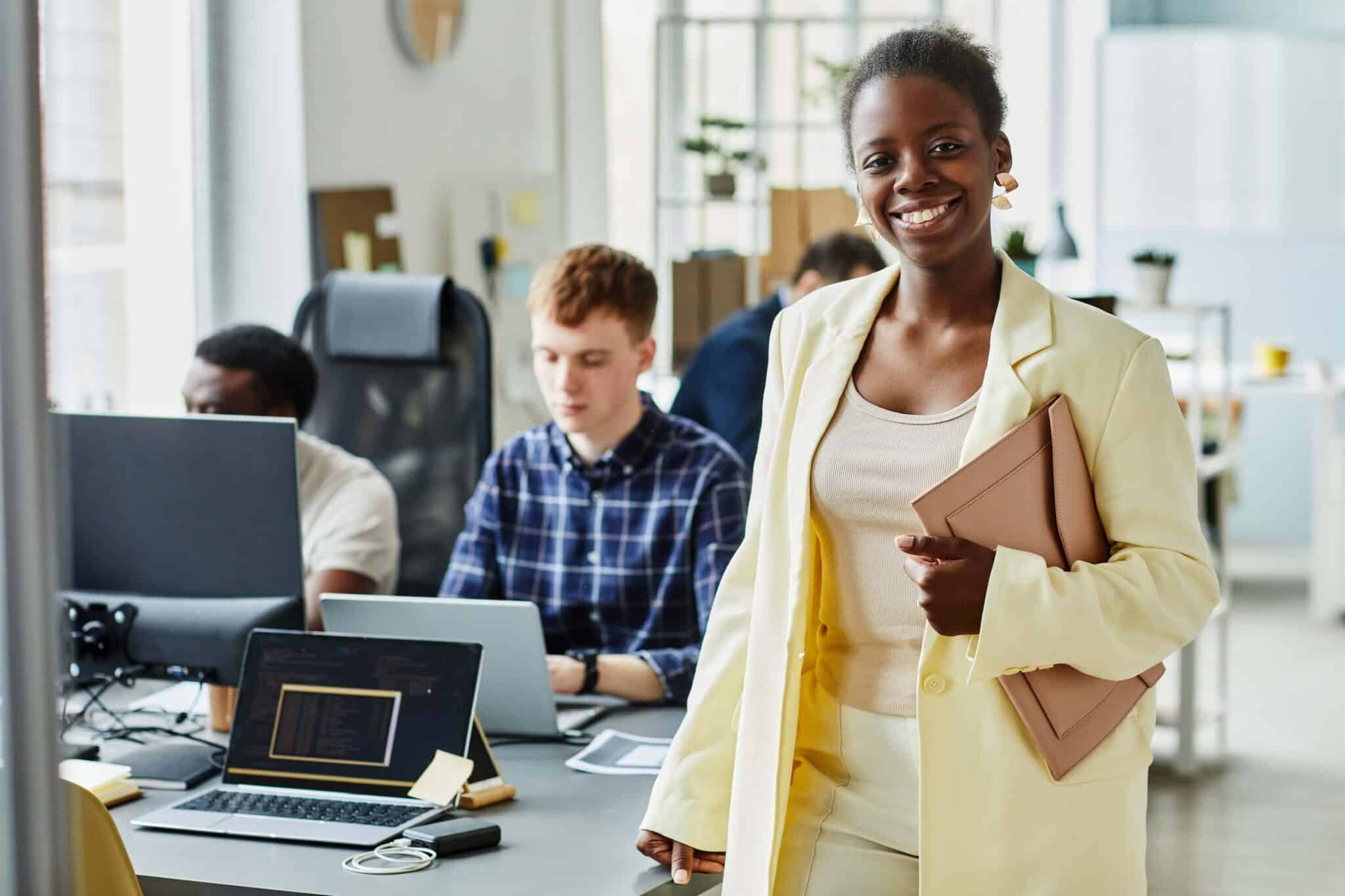 A woman in a light yellow suit smiles at the camera while holding a folder, standing in a modern office where teams discuss replicon competitors and work at desks with laptops in the background.