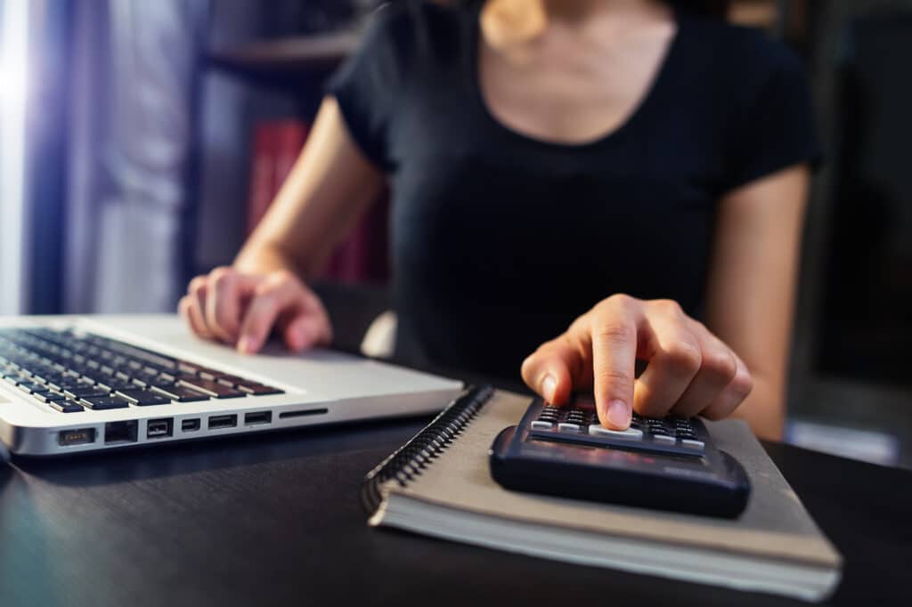 A woman wearing a black shirt uses a calculator with one hand while the other hand types on a laptop. A notebook is placed under the calculator on a dark desk. The background is blurred.