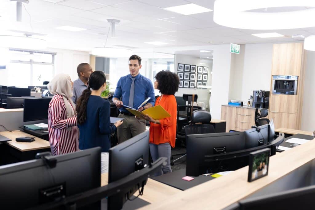 A group of six diverse coworkers are standing in a modern office, discussing project management and holding documents. Several desks and computer monitors are visible in the foreground, creating a collaborative and professional atmosphere.