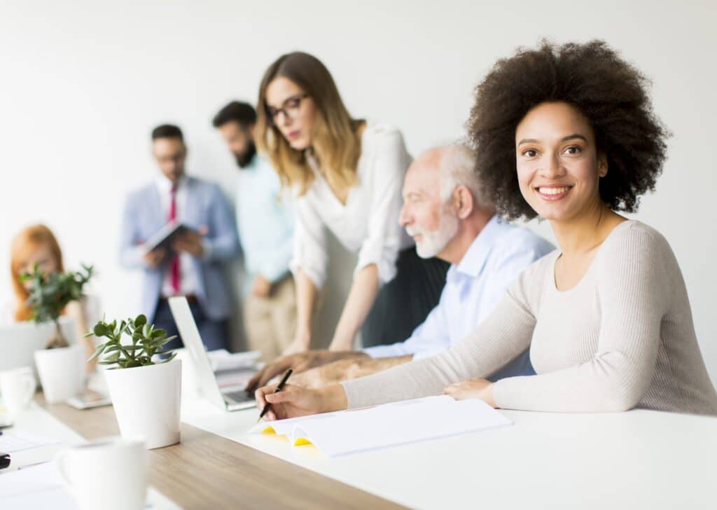 A woman smiles at the camera while sitting at a conference table with paperwork, managing time tracking. Four other colleagues are working and talking in the background of a modern, bright office.