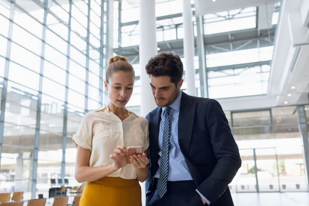 Two business professionals stand together in a modern office building, looking at a smartphone held by the woman. The man wears a suit and tie; the woman wears a blouse and skirt. Sunlight streams through large glass windows behind them.