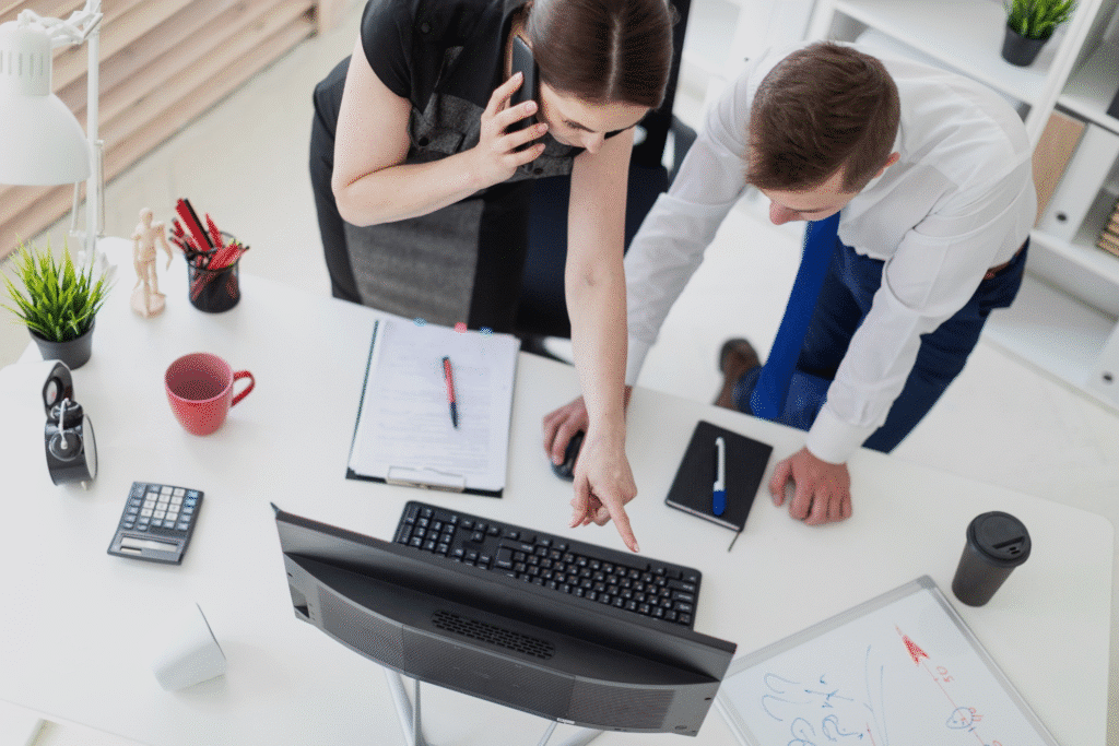 Two people in business attire are working at a desk. One is on the phone and pointing at a computer screen, possibly discussing expense coding, while the other looks on. Office supplies, a keyboard, and paperwork are visible on the white desk.