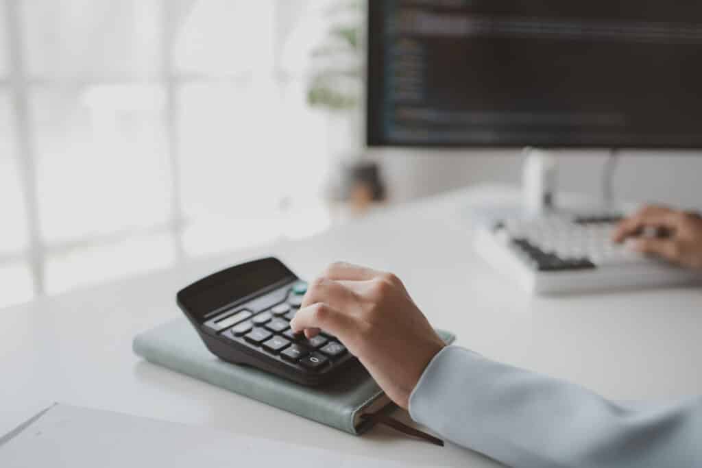 A person uses a calculator on a notebook while working at a desk, with another hand on a computer keyboard and a monitor displaying code in the background.