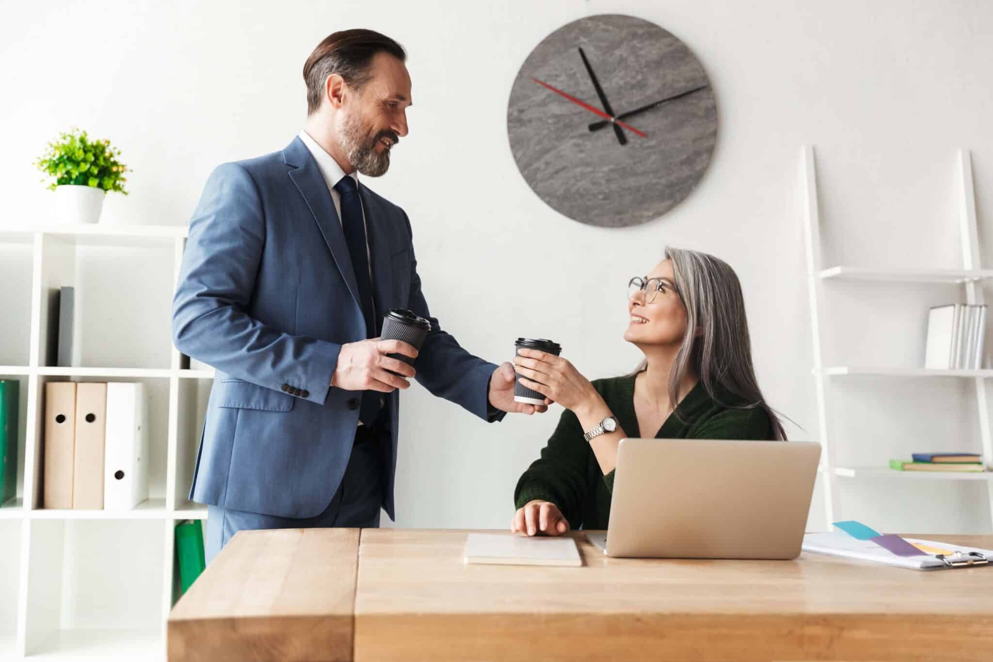 A man in a suit hands a coffee cup to a smiling woman with gray hair sitting at a desk with a laptop, showcasing teamwork in a modern office featuring a large wall clock—an ideal setting for using the best time and attendance software.