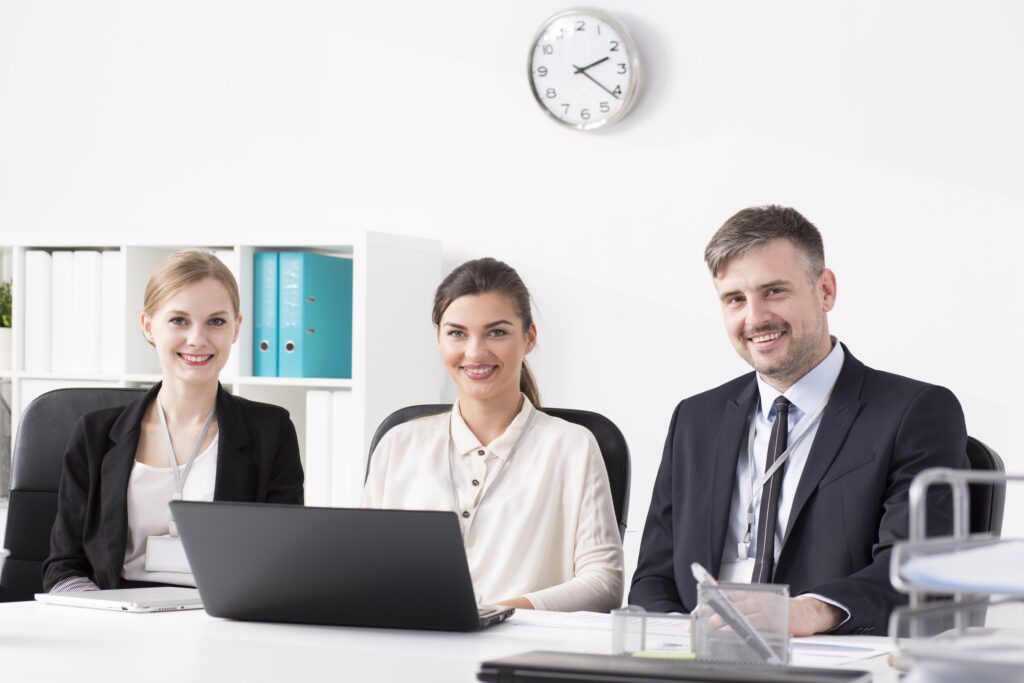 Three professionally dressed people sit at a desk in an office, smiling at the camera. A laptop—perhaps used for timesheet automation—is open in front of them, with shelves of binders and a clock on the white wall showing 10:10.