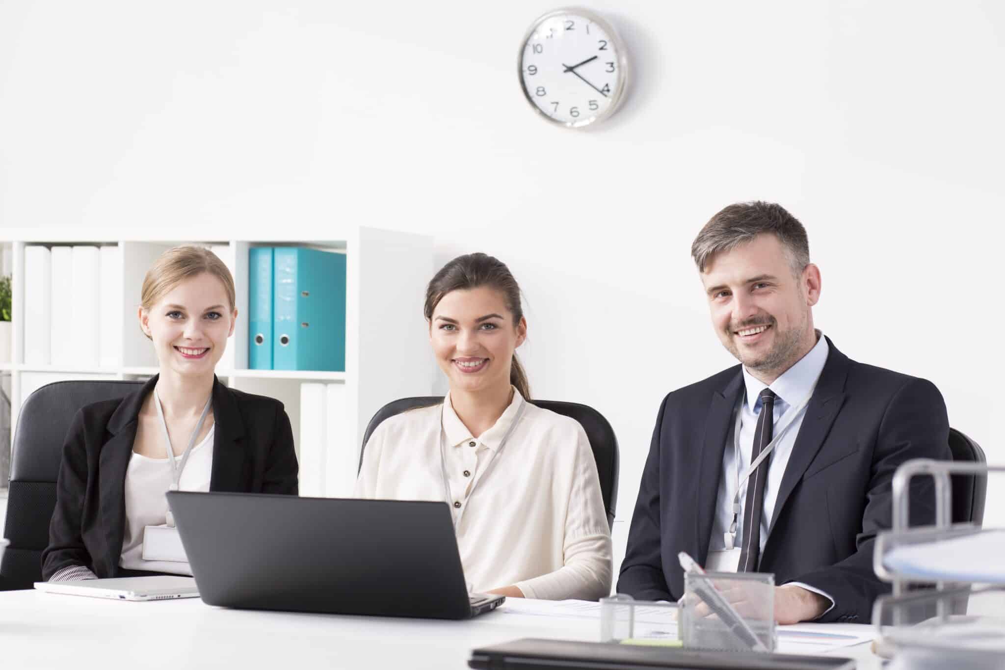 Three professionally dressed people sit at a desk in an office, smiling at the camera. A laptop—perhaps used for timesheet automation—is open in front of them, with shelves of binders and a clock on the white wall showing 10:10.