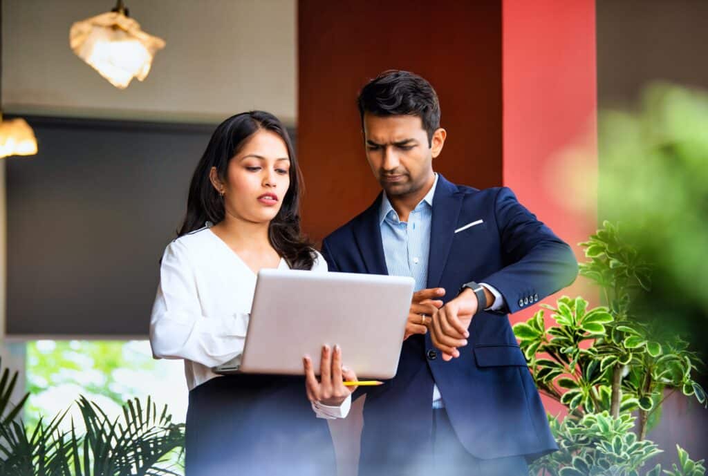 A woman in business attire holds a laptop while a man in a suit checks his watch. Surrounded by green plants, they appear focused and engaged in a project management discussion indoors.