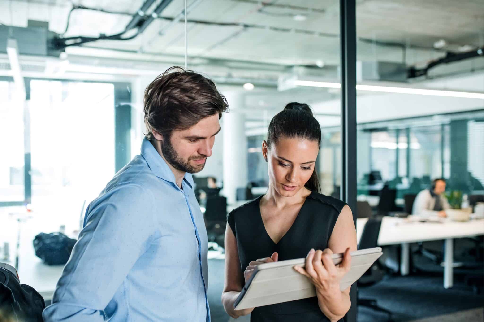 A man and a woman stand together in a modern office, looking at a tablet the woman is holding. Both appear focused and engaged in discussion about timesheet compliance. Open workspace and coworkers are visible in the background.