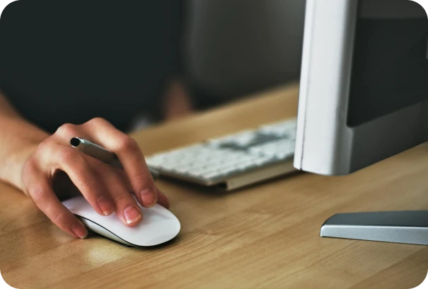 A person’s hand holding a pen while using a computer mouse, with a keyboard and monitor on a wooden desk.