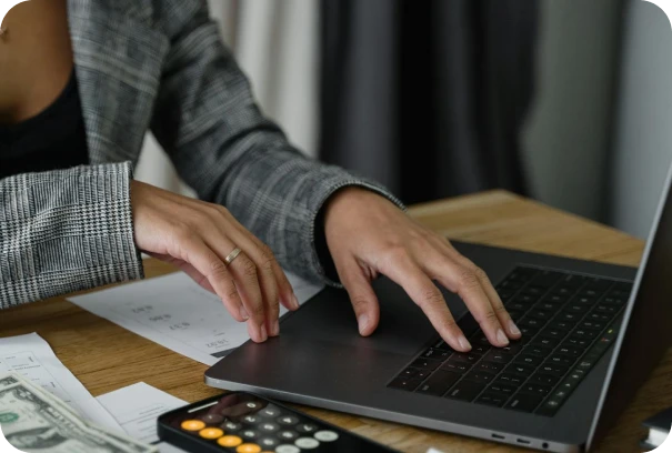 A person in a plaid blazer types on a laptop at a wooden desk with a calculator, US dollar bills, and documents nearby.