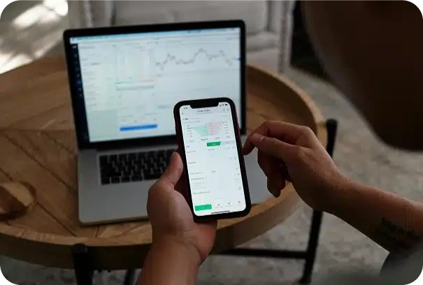 A person checks financial or stock trading data on a smartphone while a laptop displaying a chart and data is open on a wooden table in the background.