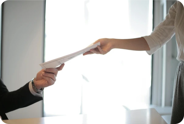 A person in business attire hands documents to another person across a desk in a bright office setting.