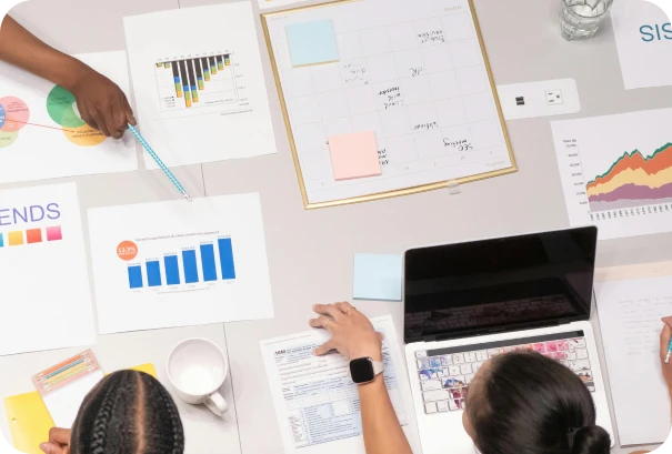 Three people sit at a table with charts, graphs, sticky notes, and a laptop, collaborating and reviewing data and paperwork in a meeting or brainstorming session (time billing and invoicing software).
