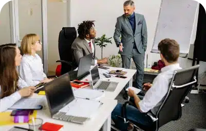 Four people sit at a conference table with laptops and notebooks while a man in a gray suit stands and gestures toward them, presenting information on a flip chart in an office setting.