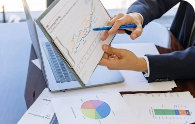 A person in a suit points to a clipboard with a pen, showing a graph with rising trends, while sitting at a desk with a laptop and various financial charts and documents.