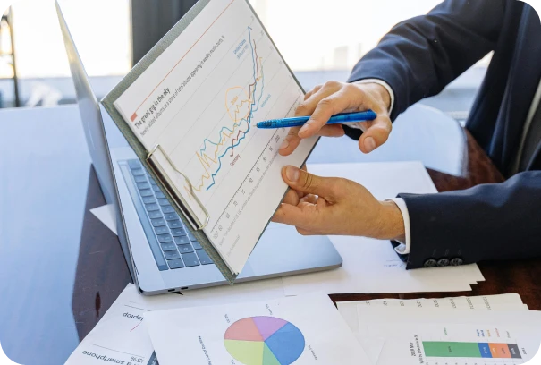 A person in business attire points at a financial chart on a clipboard, showing line graphs, while sitting at a desk with a laptop and papers displaying pie and bar charts.