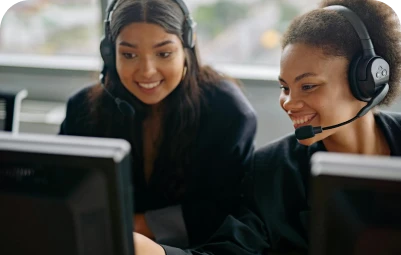 Two women wearing headsets sit side by side at computer monitors, smiling and looking at the screens, suggesting they are working together in a customer service or call center environment.
