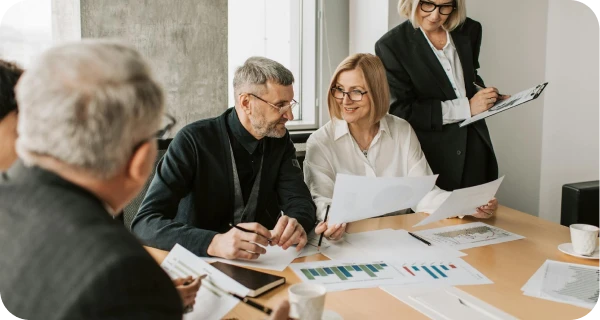 Four professionals sit around a conference table reviewing charts and papers, while a fifth person stands and takes notes on a clipboard in a bright office setting.