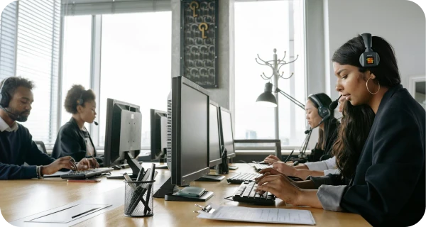 Four people wearing headsets work at computers in a modern office with large windows. They appear focused on their tasks, suggesting a call center or customer service environment.