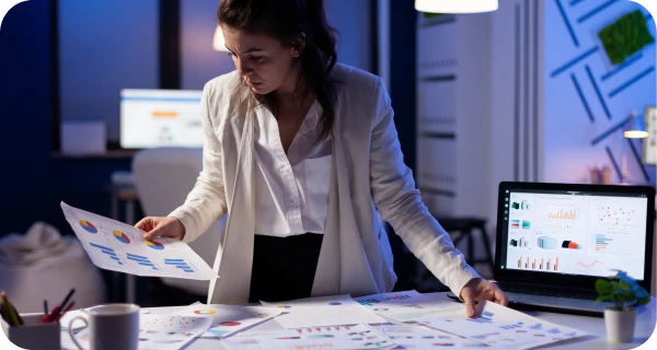 A woman in a white blazer reviews colorful charts and graphs spread across a desk in a dimly lit office, with a laptop displaying data visualizations beside her.