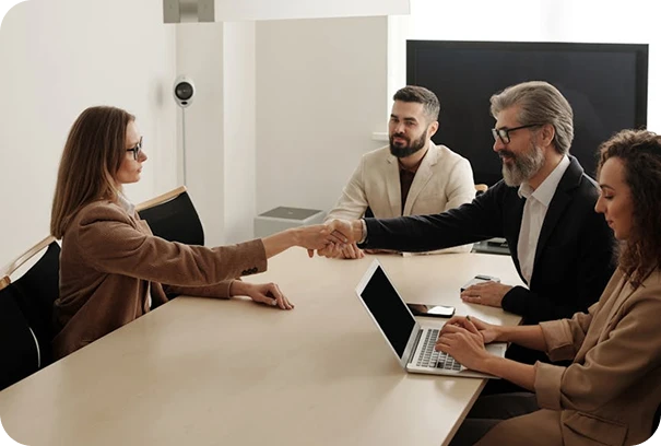 Four people sit at a conference table; one woman is shaking hands with a man across from her. Two others, a man and a woman with a laptop, observe the interaction, suggesting a business meeting or job interview.