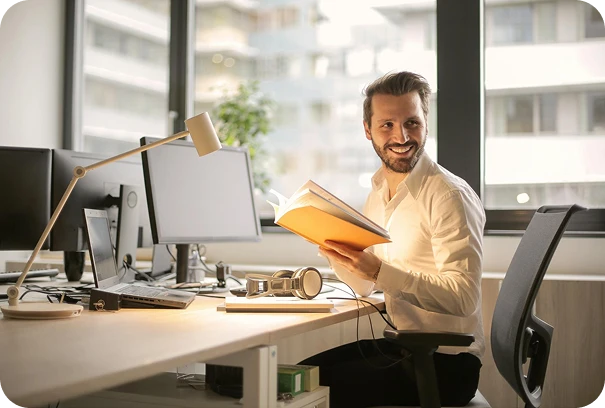A man in a white shirt sits at a desk with computers, smiling while holding an open book. Sunlight streams through large windows behind him, creating a bright office environment.