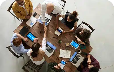 A group of people sitting around a wooden table with laptops and notebooks, seen from above. Two people in the center are shaking hands, indicating a collaborative meeting or agreement.