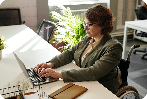 A person in a wheelchair, wearing glasses and a blazer, types on a laptop at a desk in a bright office with plants and sunlight streaming in. A notebook is placed on the desk nearby.