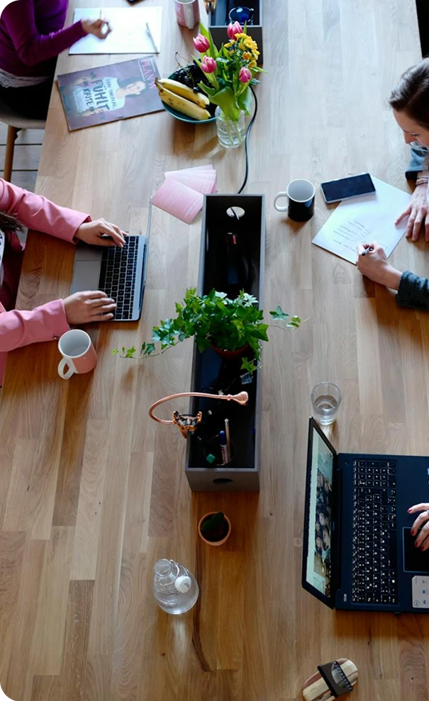 Overhead view of a wooden table with laptops, notebooks, coffee mugs, and plants. Three people are working, one typing, another writing notes, and a vase of flowers and fruit bowl are also visible.