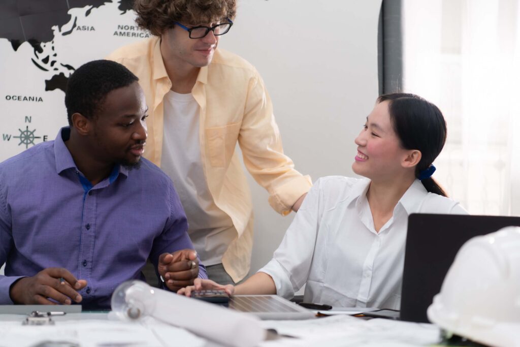 Three colleagues, two men and one woman, sit and stand around a desk with a laptop, papers, and a hard hat, smiling and engaging in conversation. A world map is visible in the background.