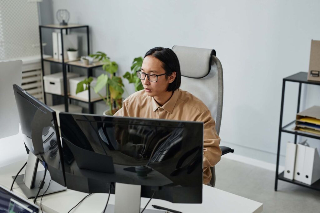 Person with glasses and shoulder-length hair sits at a desk working on a computer with dual monitors in a modern office, surrounded by shelves with folders, plants, and office supplies.