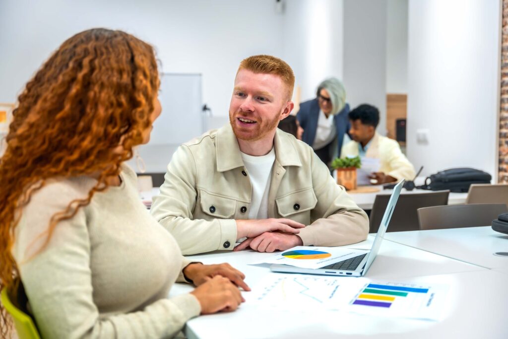 Two people sit at a table discussing documents with charts, a laptop open in front of them, while others work in the background in a modern, bright office environment.