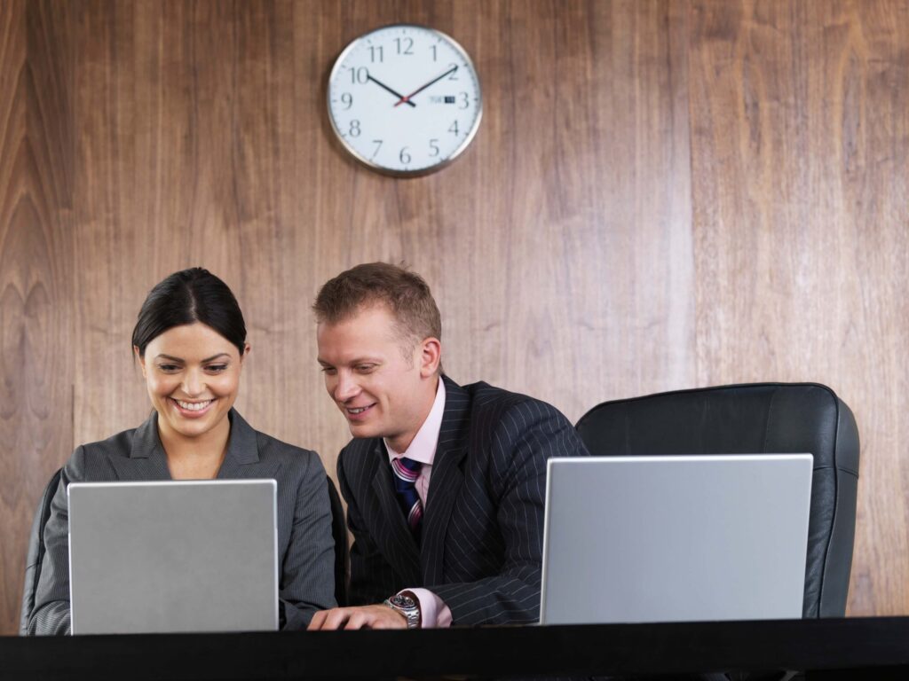 Two business professionals, a woman and a man in suits, are smiling and looking at a laptop screen together at a desk with another laptop—perhaps discussing how to get employees to submit timesheets on time before the pay period ends. A wall clock shows 9:16 behind them.