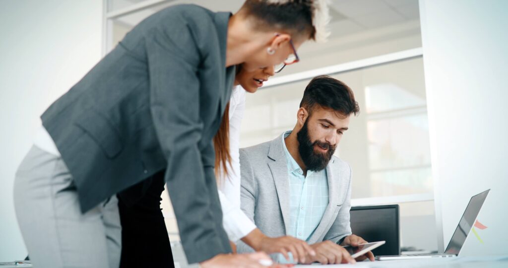 Three business professionals collaborate at a desk, reviewing documents and using a tablet. One man is seated, while two women stand beside him, all focused and engaged in discussion in a bright office setting.