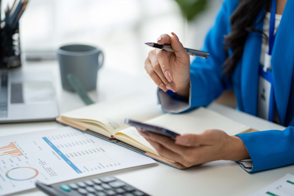 A person in a blue blazer sits at a desk holding a pen and smartphone, with an open notebook, a report with charts, a coffee mug, and a calculator in view.