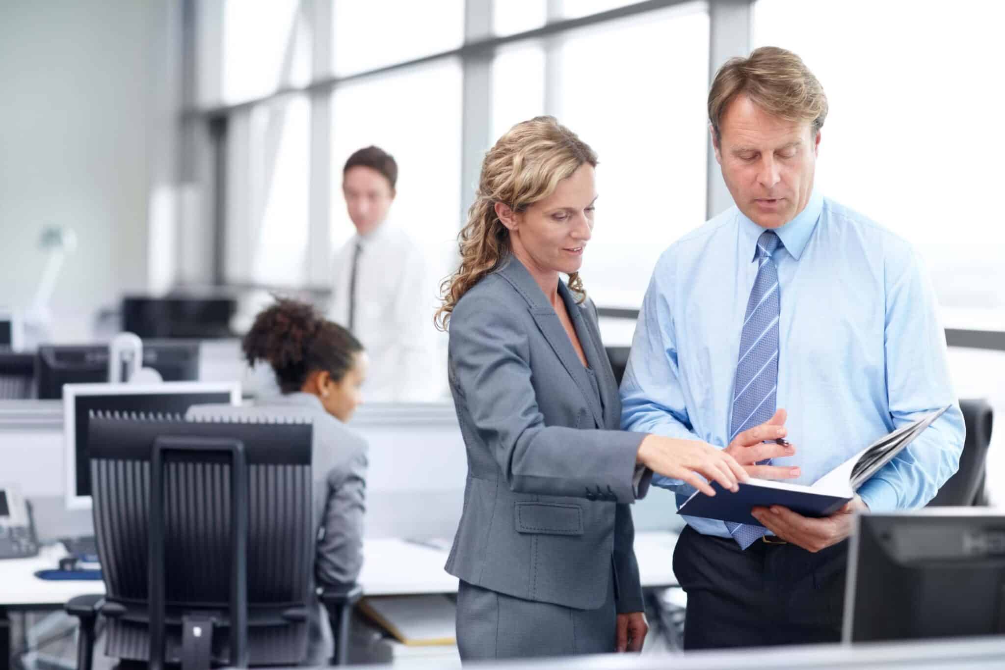 Two business professionals in suits stand in an office, reviewing an invoice discrepancy together. Other colleagues work at desks in the background. The setting is bright with large windows.